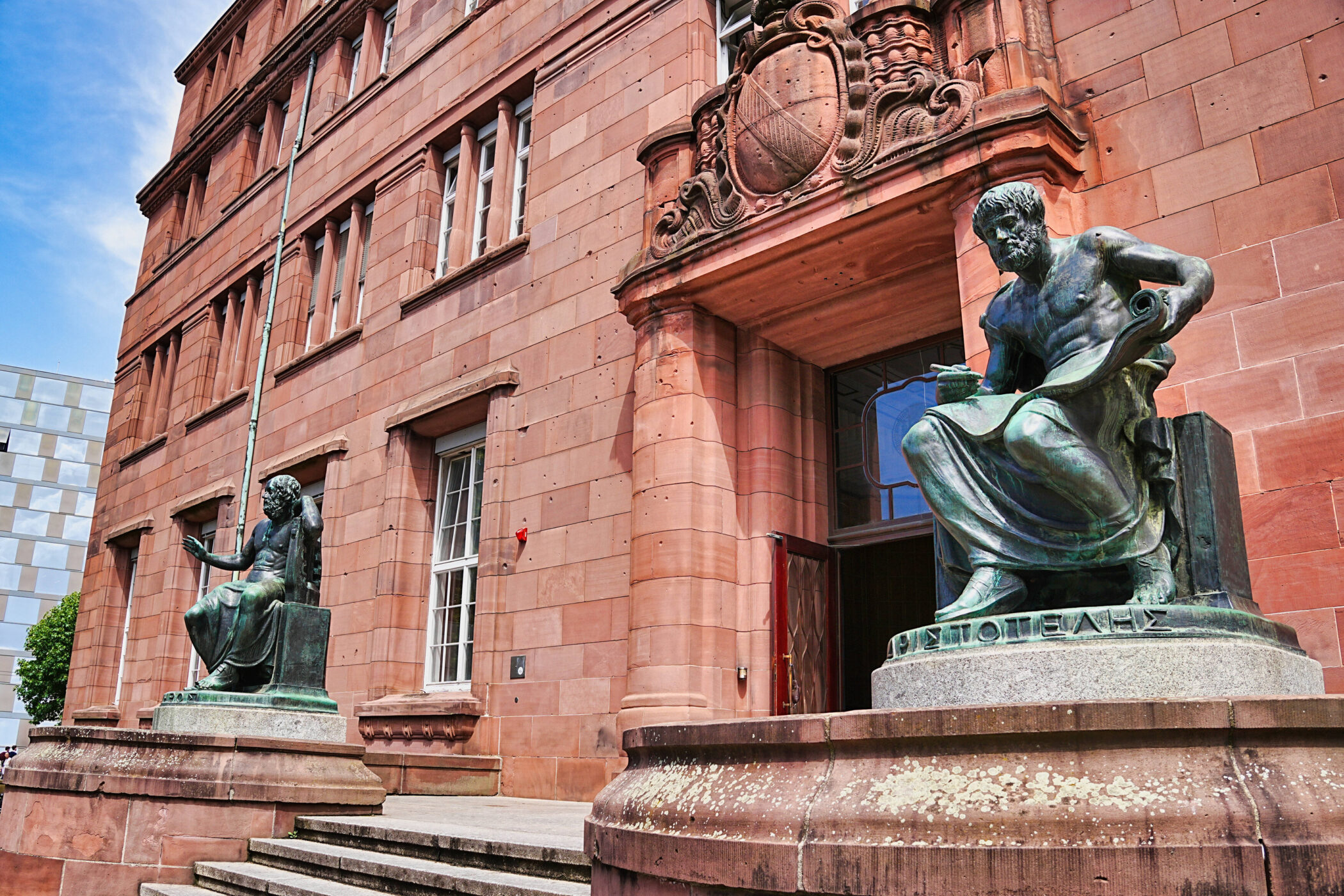 Zwei Bronzestatuen vor einem roten Gebäude mit vielen Fenstern.