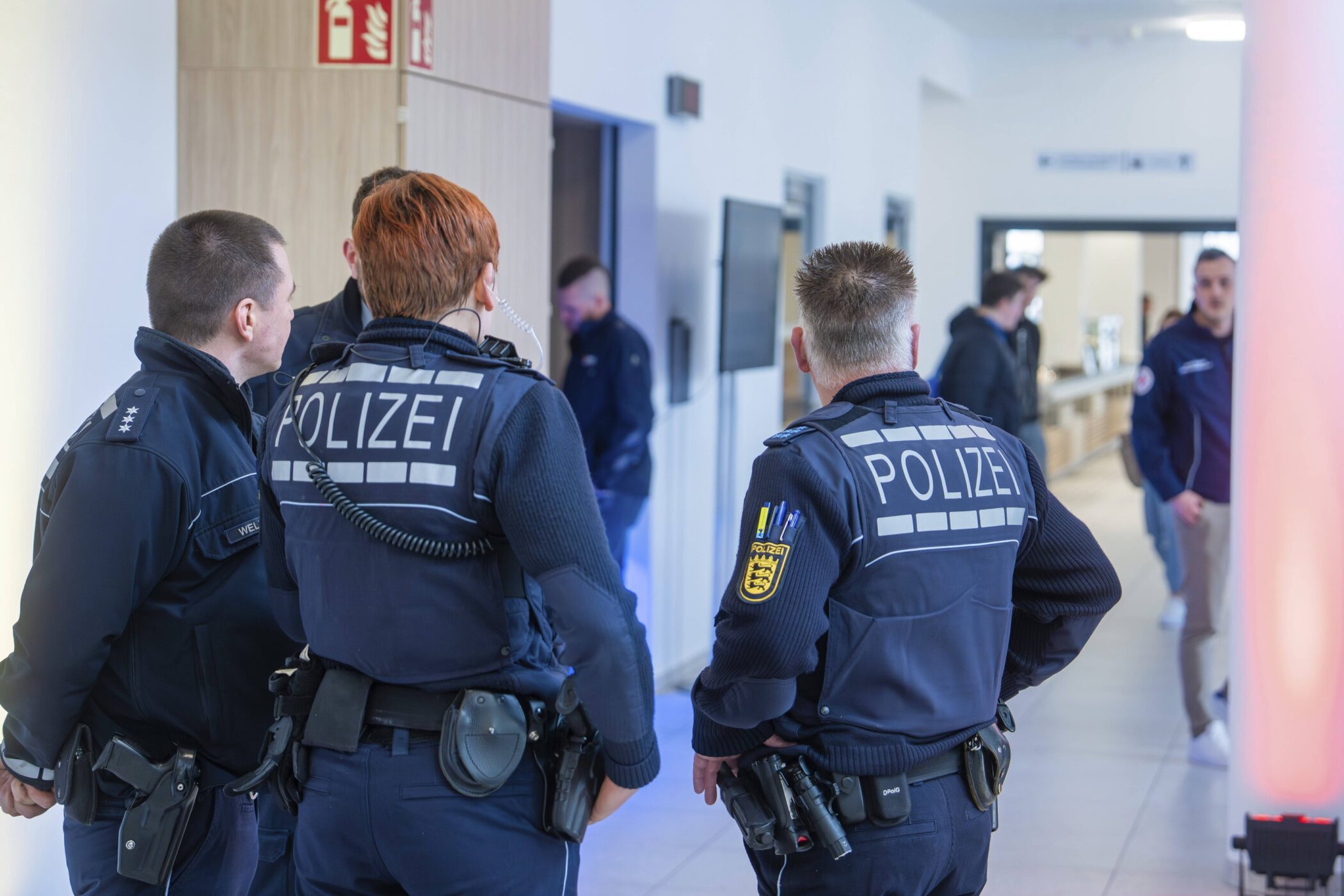 Drei Polizisten von hinten in einem Flur, mehrere Personen im Hintergrund.