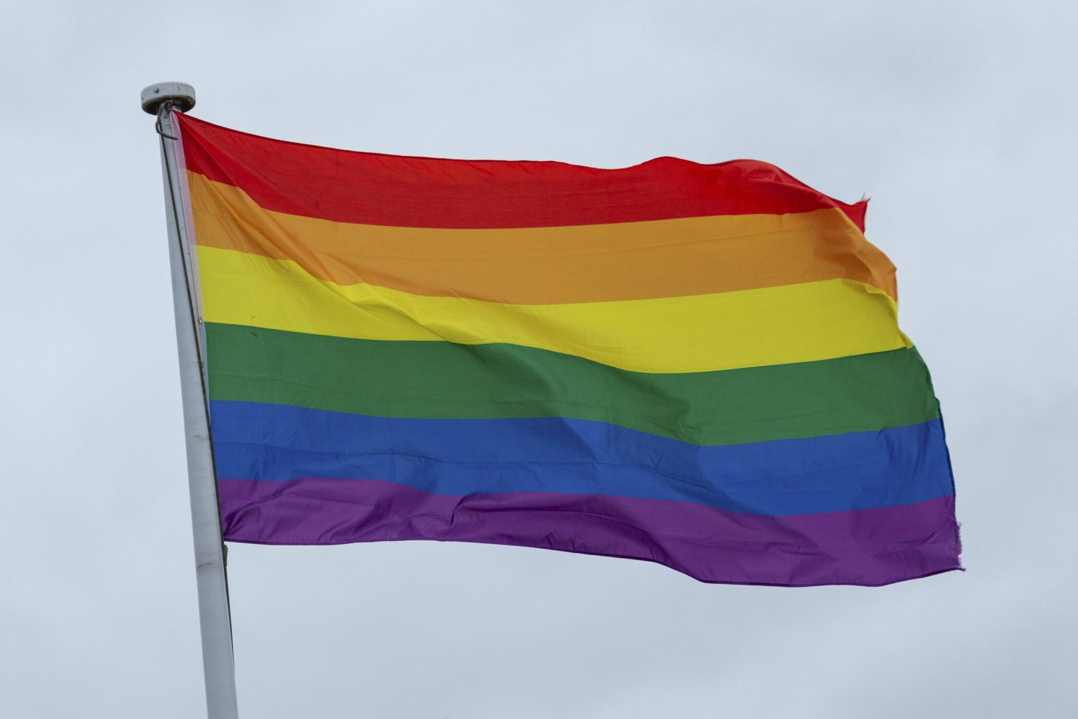 Regenbogenflagge weht an einem Fahnenmast vor bewölktem Himmel.