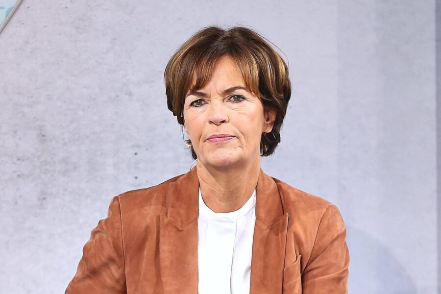 Eine Frau in brauner Jacke und weißem Oberteil vor grauem Hintergrund.