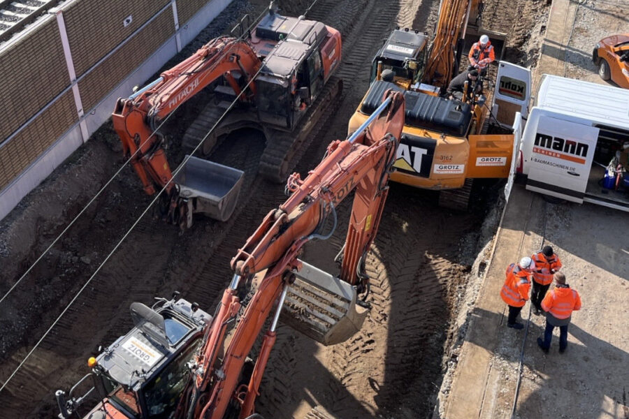 Bagger und Arbeiter auf Baustelle, Personen in Warnwesten stehen zusammen.