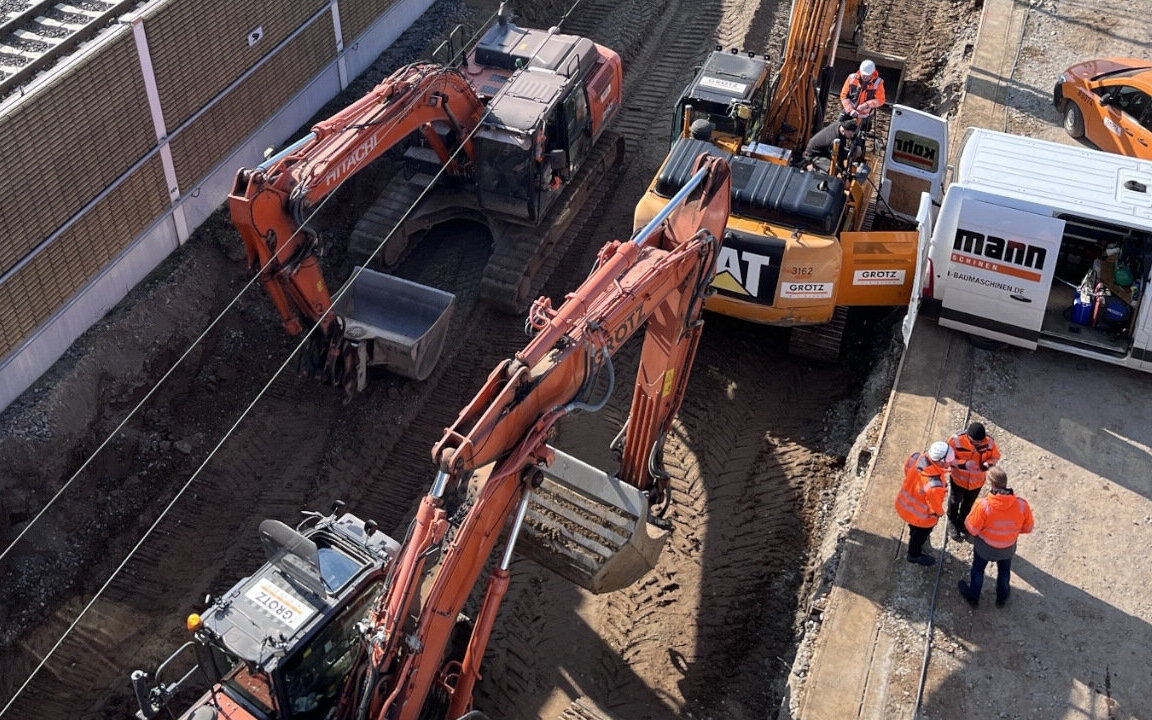 Bagger und Arbeiter auf Baustelle, Personen in Warnwesten stehen zusammen.