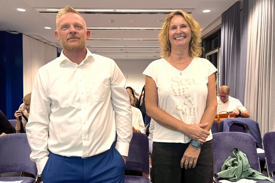 Zwei stehende Personen in einem Raum, Text auf T-Shirt: „Sea, Air & Sun“.