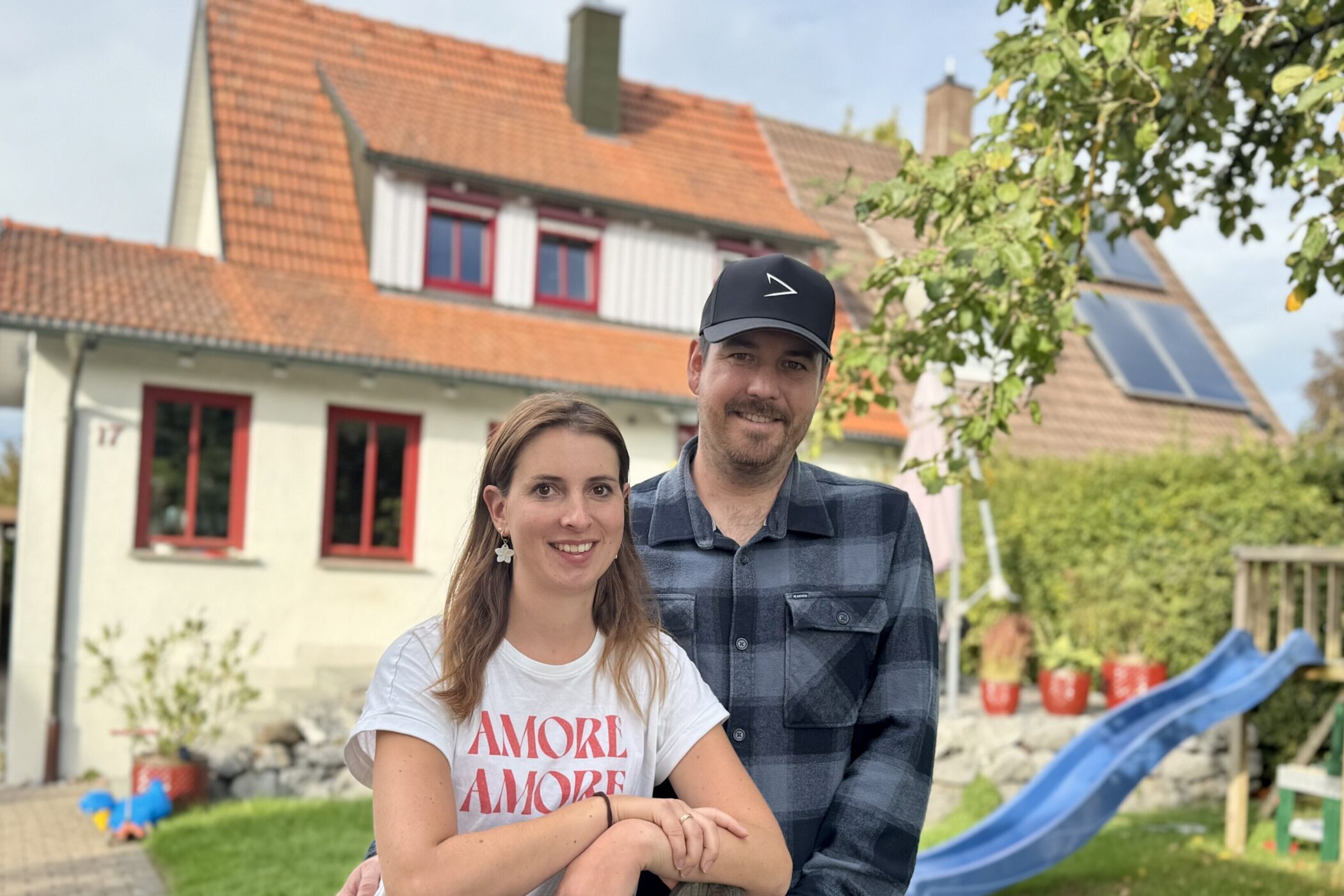 Zwei Personen stehen lächelnd vor einem Haus mit roten Fenstern und Gartenrutsche.