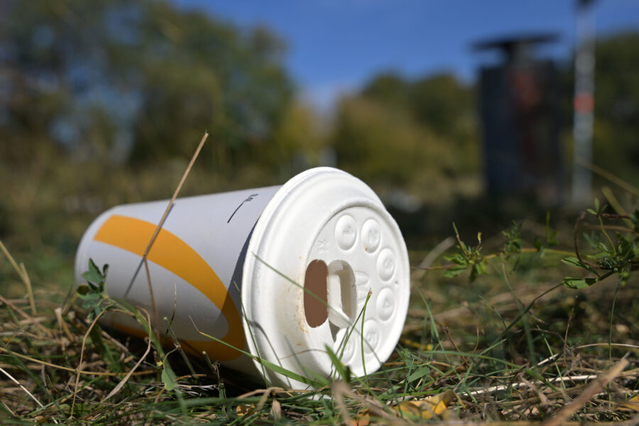 Papierbecher mit Deckel liegt auf Gras, im Hintergrund Bäume.