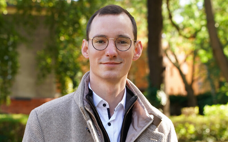 Ein Mann mit Brille in Jacke steht draußen vor unscharfem Hintergrund.