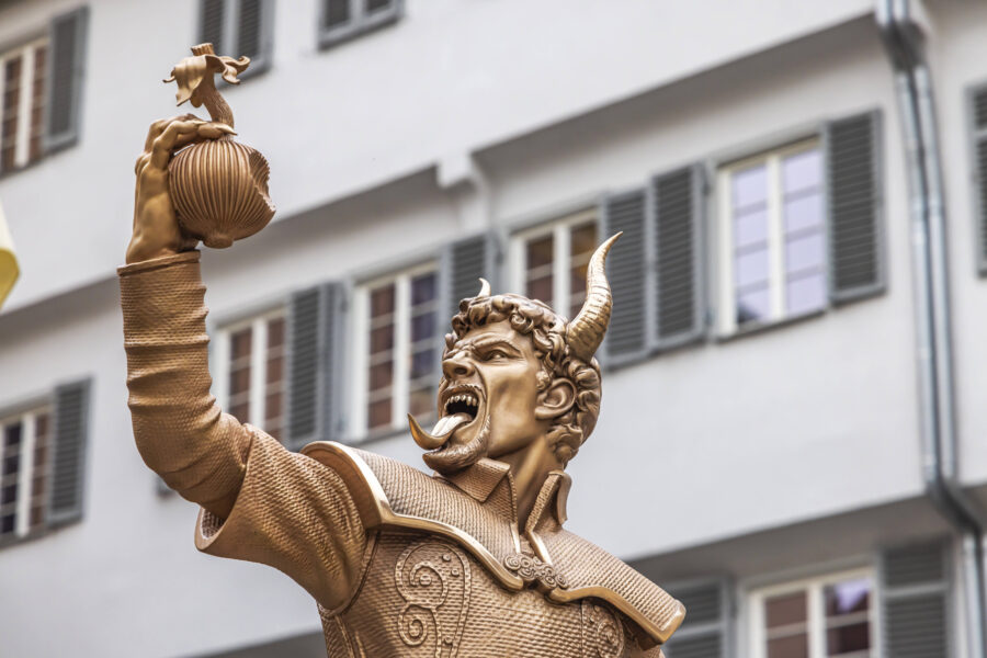 Goldene Statue eines Mannes mit Hörnern, hält einen Fußball, Gebäude im Hintergrund.