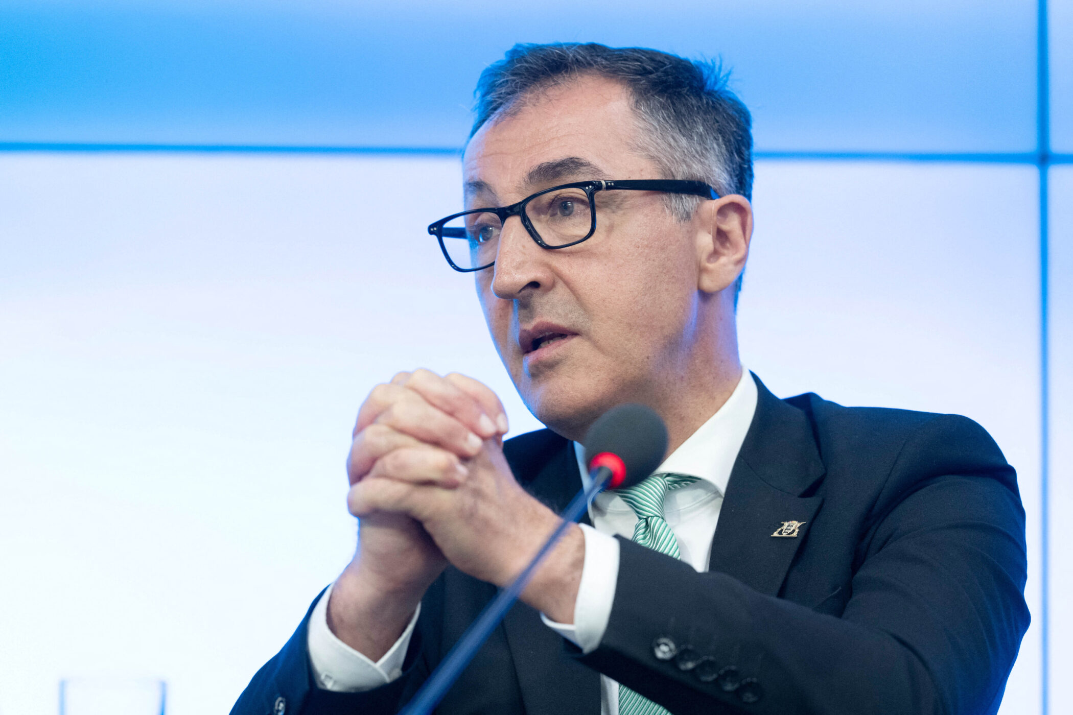 Mann mit Brille, gestikuliert, Mikrofon im Vordergrund, blauer Hintergrund.