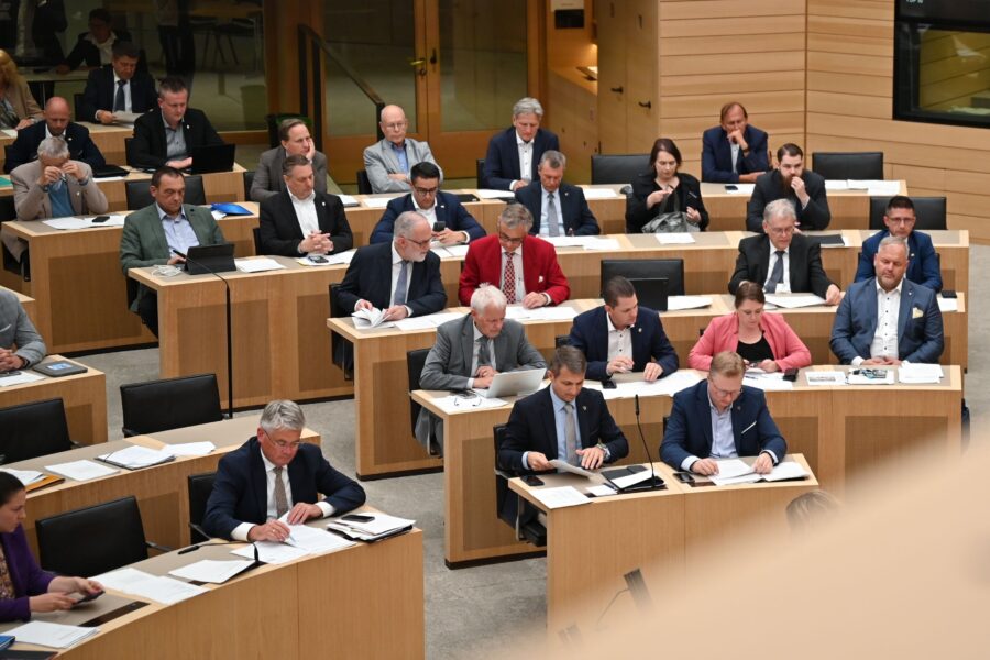 Mehrere Personen in einem Sitzungssaal an Holztischen, einige mit Laptops.