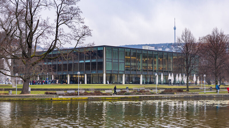 Rechteckiges Gebäude mit Glasfassade, Bäume, Teich im Vordergrund, Fernsehturm im Hintergrund.
