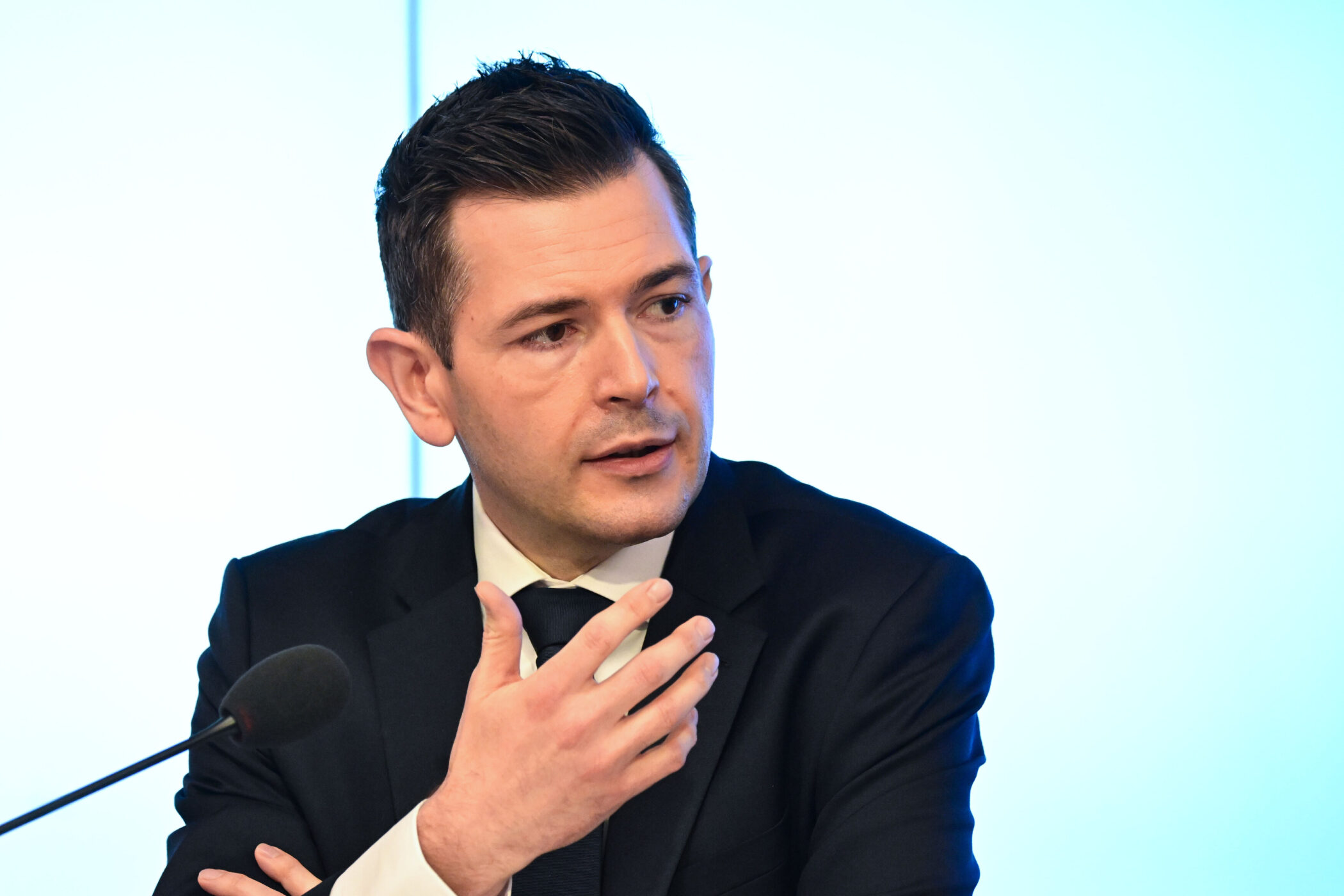 Mann im Anzug spricht, Mikrofon vor ihm, blauer Hintergrund.
