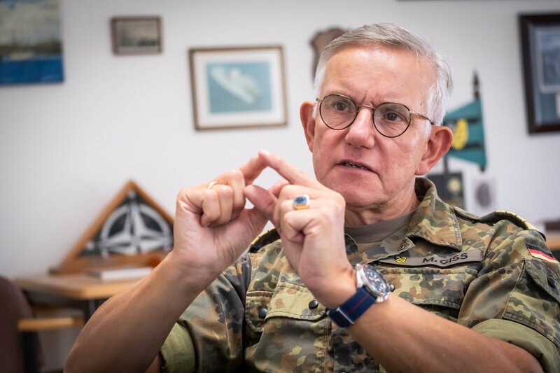 Älterer Mann in Uniform spricht gestikulierend, Wände mit gerahmten Bildern.