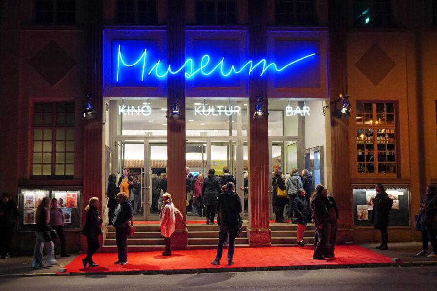 Vor einem Gebäude mit der Aufschrift „Museum“ stehen mehrere Menschen auf rotem Teppich.