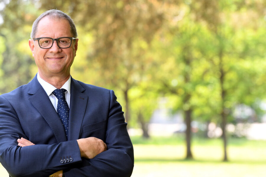 Mann im Anzug mit Brille, Arme verschränkt, Hintergrund Bäume und Wiese.