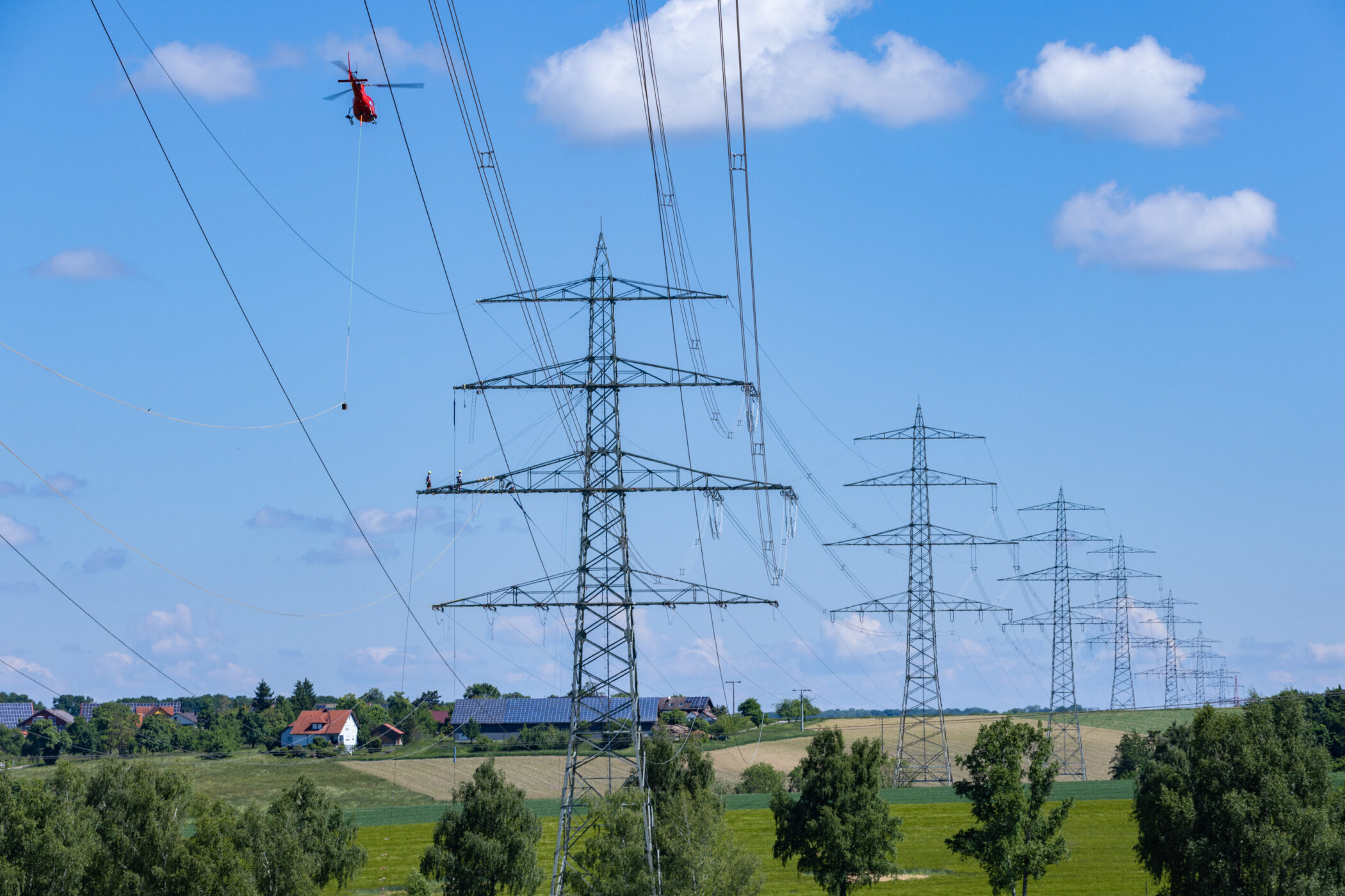 Roter Helikopter fliegt nahe an Strommasten über grüner Landschaft mit Häusern.