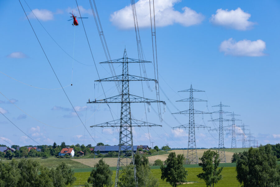 Roter Helikopter fliegt nahe an Strommasten über grüner Landschaft mit Häusern.