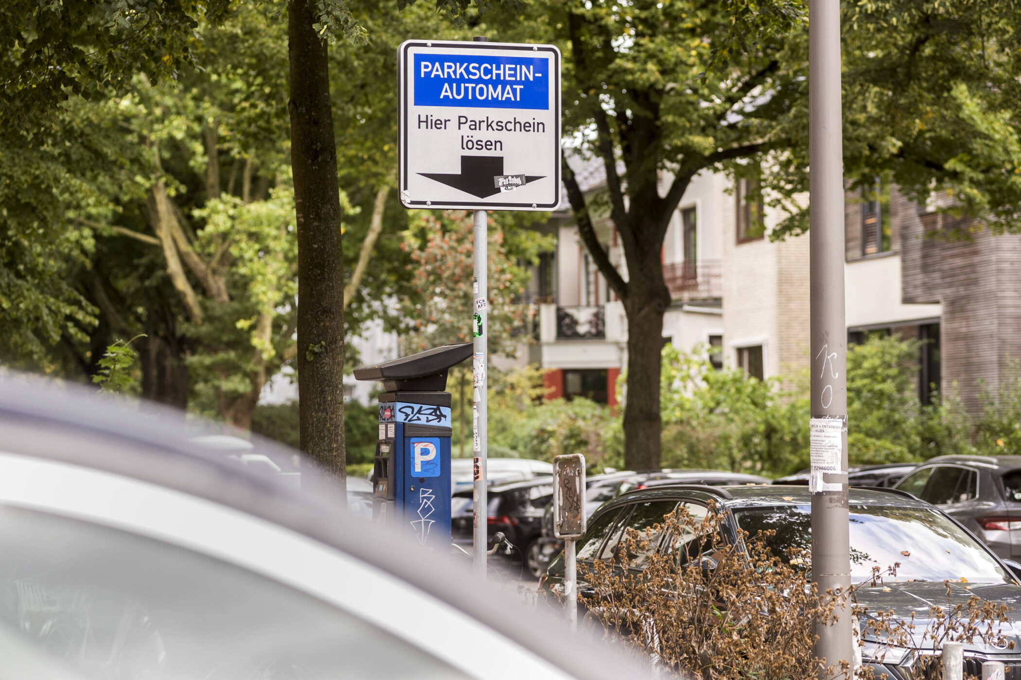Parkscheinautomat und Schild "Hier Parkschein lösen" in einer Baum gesäumten Straße.