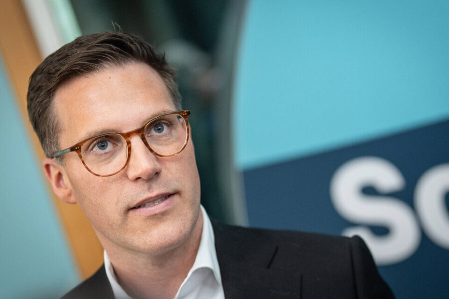 Person mit Brille vor blauer Fläche, Text "Sc..." sichtbar.
