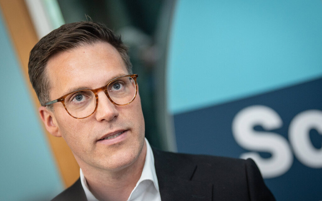 Person mit Brille vor blauer Fläche, Text "Sc..." sichtbar.