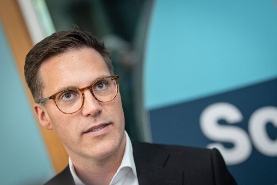 Ein Mann mit Brille vor einem Schild mit unleserlichem Text.