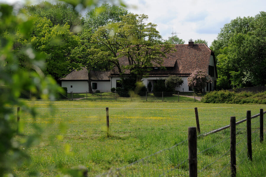 Weißes Haus mit Garten, umgeben von Bäumen, im Hintergrund eines grünen Feldes.