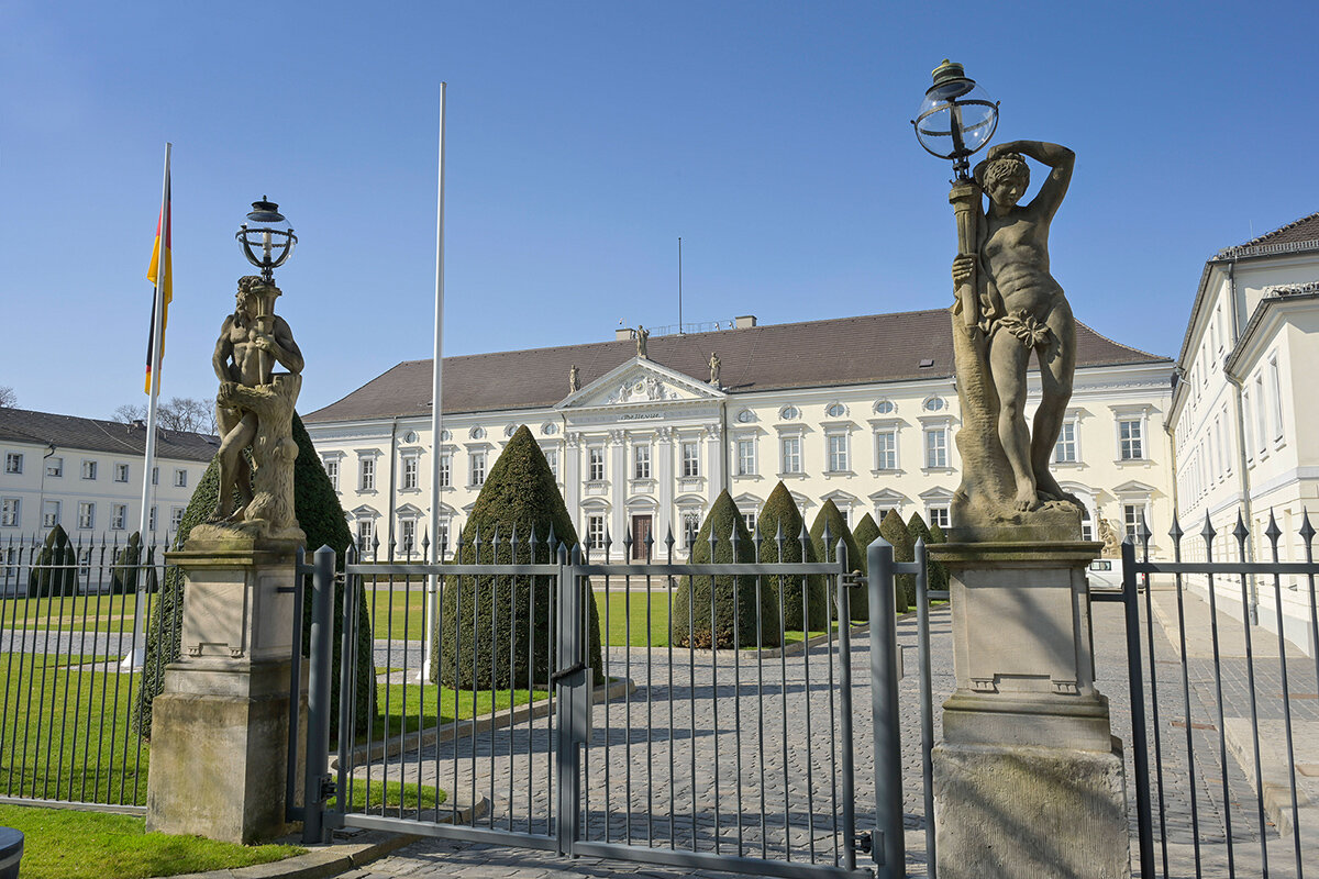 Schloss mit zwei Skulpturen am Tor, deutsche Flagge links, geschnittener Baum.