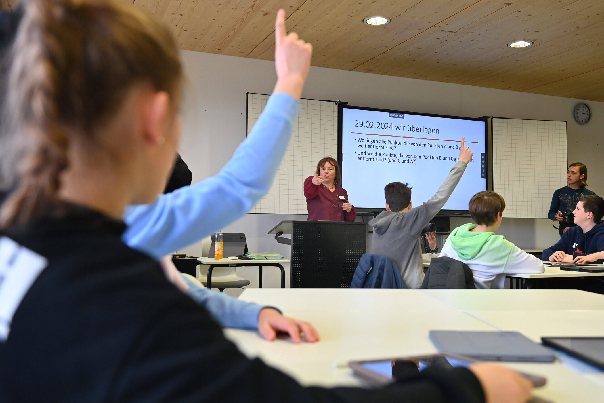 Mehrere Schüler in einem Klassenzimmer heben die Hand, eine Frau zeigt auf sie.