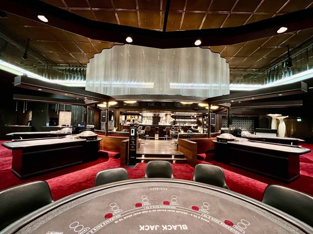 Großer Raum mit Blackjack-Tisch, umgeben von weiteren Spieltischen und Bar im Hintergrund.