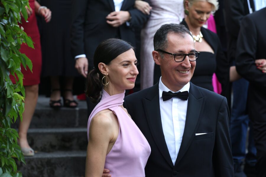 Eine Frau im rosa Kleid und ein Mann im Anzug lächeln, Menschen im Hintergrund.