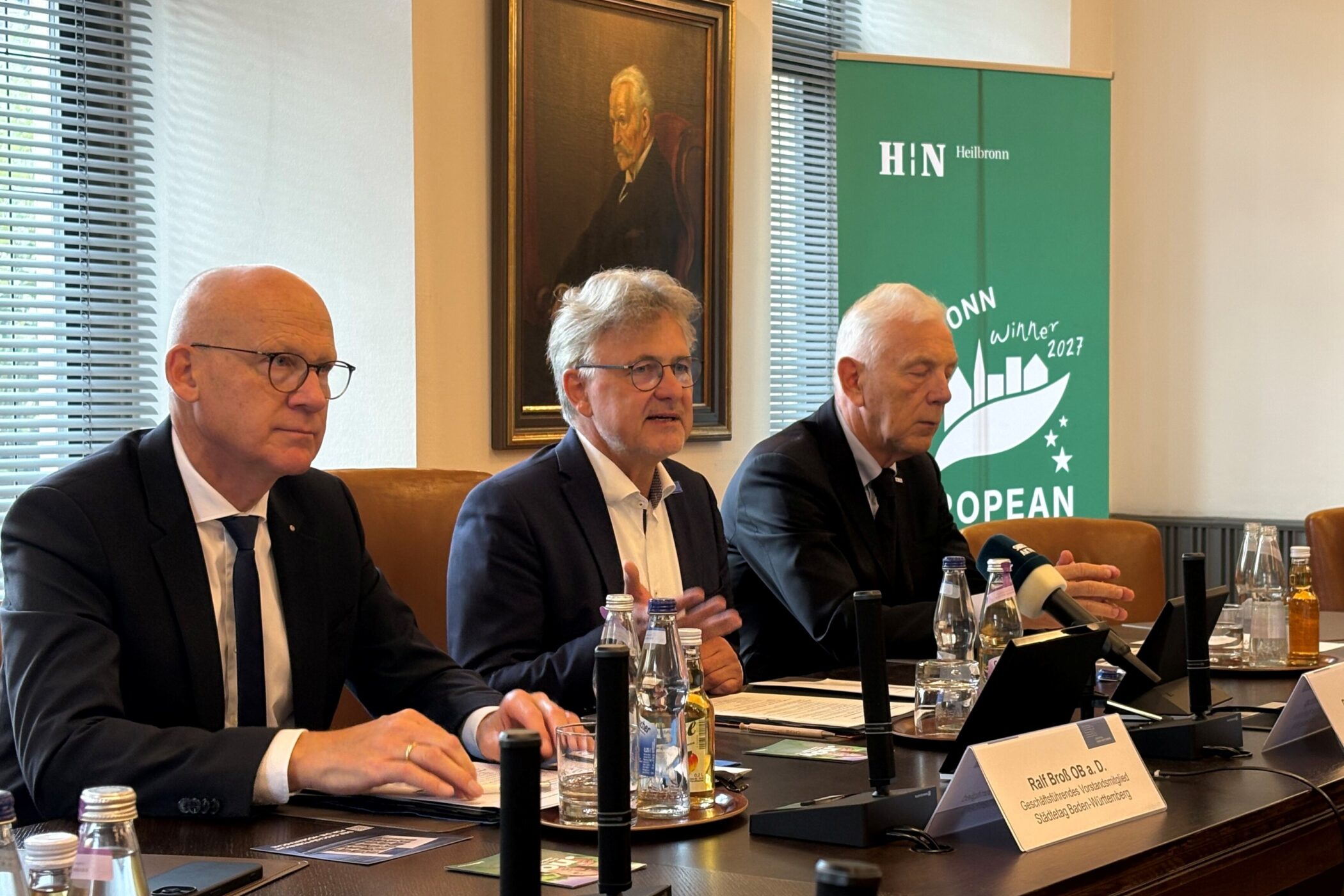 Drei Männer im Anzug sitzen an einem Konferenztisch mit Mikrofonen.