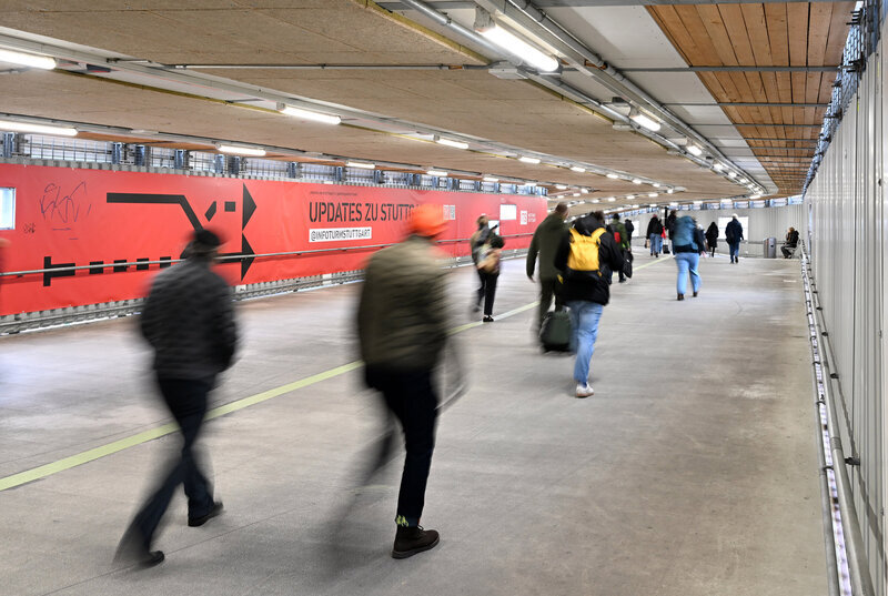 Personen gehen durch einen Tunnel, rote Werbetafel rechts mit Text.