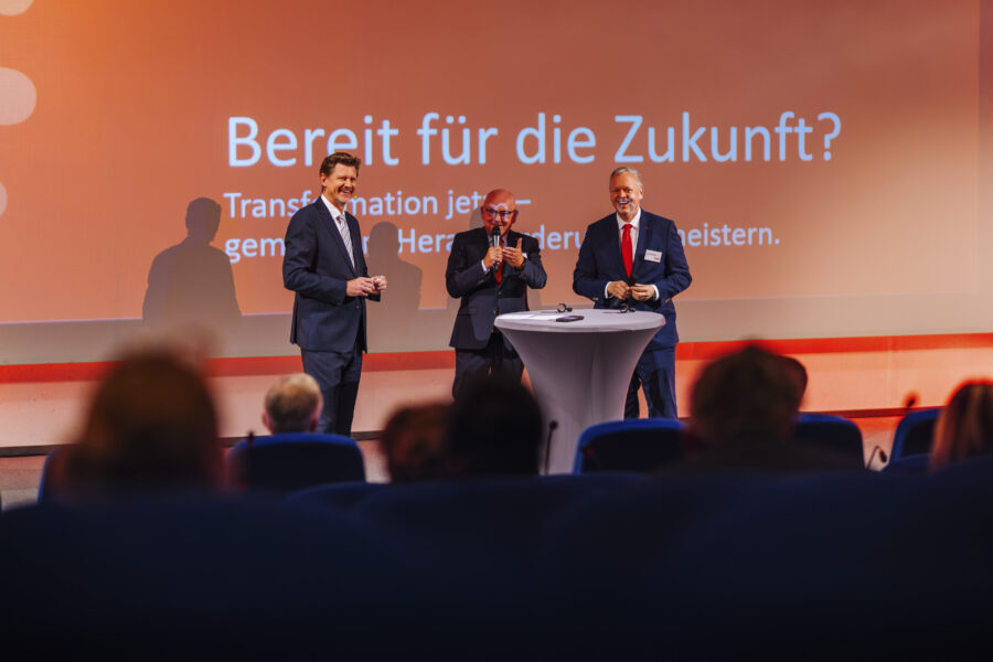 Drei Männer in Anzügen auf Bühne, Text im Hintergrund: "Bereit für die Zukunft?"