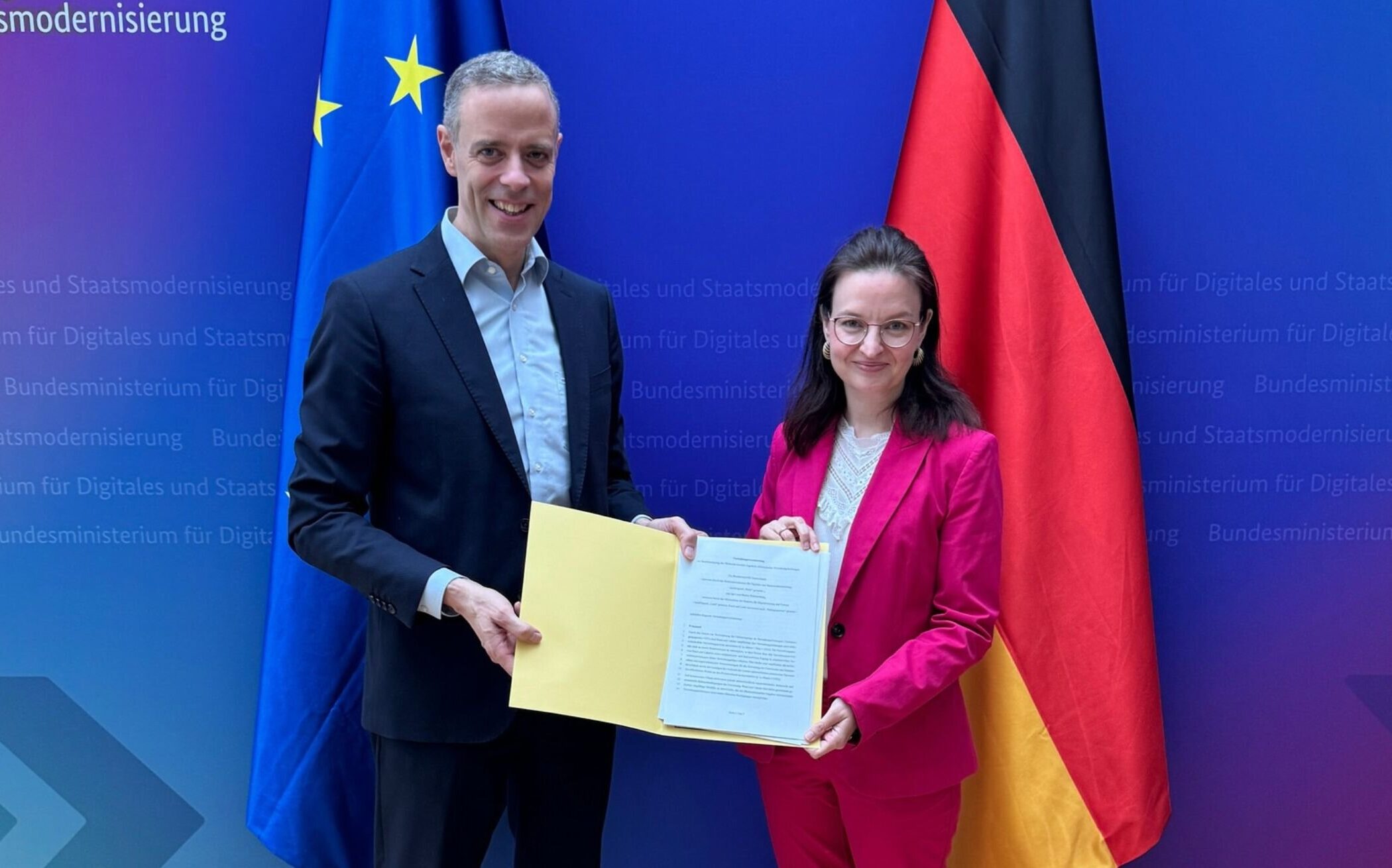 Zwei Personen halten ein Dokument vor EU- und Deutschlandflagge. Hintergrund: Ministeriumswand.