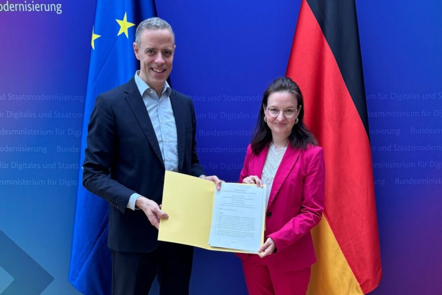 Zwei Personen halten ein Dokument vor EU- und Deutschlandflagge. Hintergrund: Ministeriumswand.