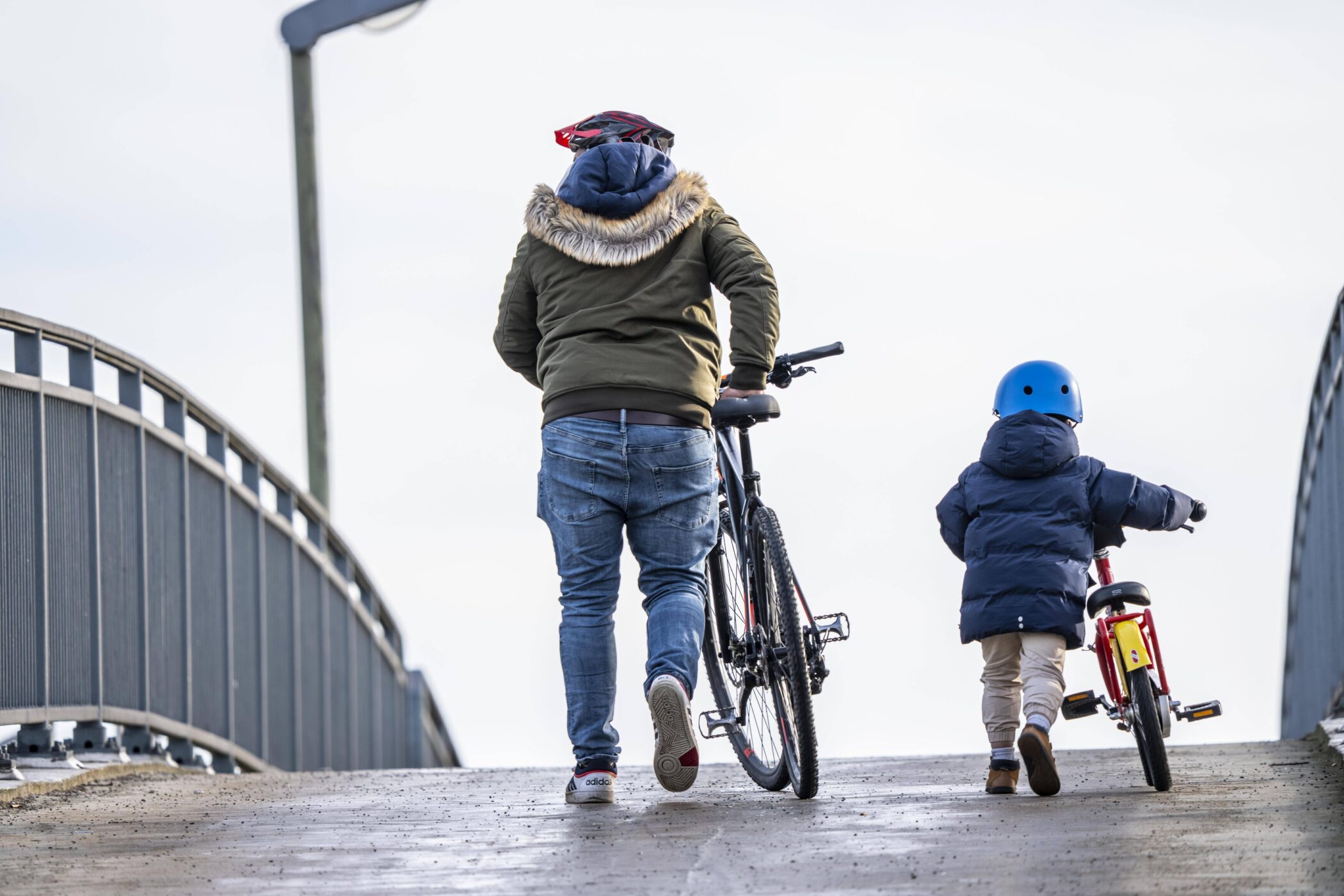 Eine Person mit Fahrrad, ein Kind mit Laufrad auf einer Brücke von hinten.