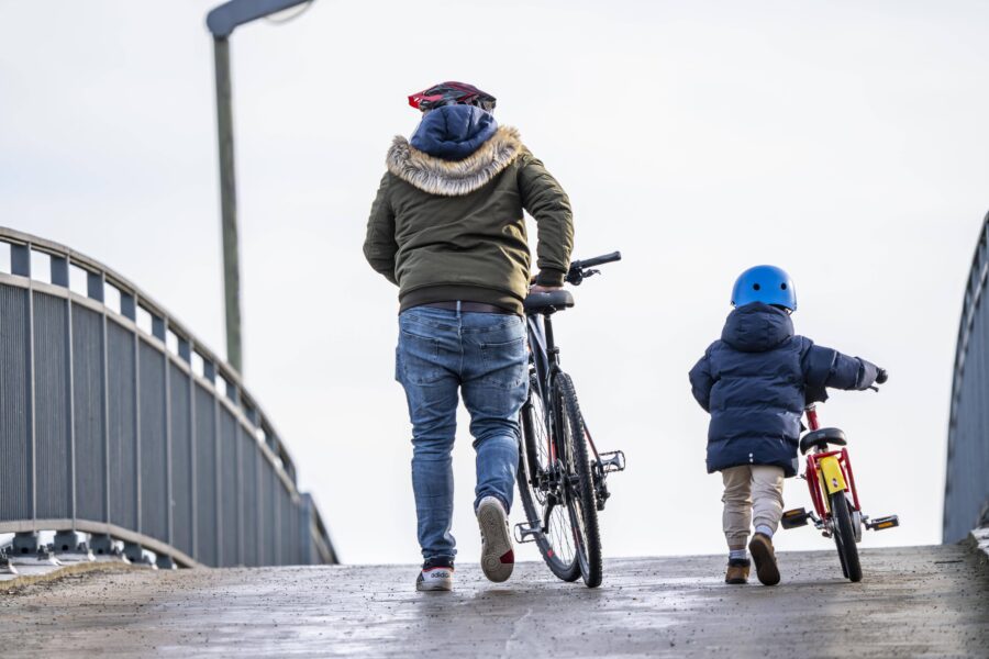 Eine Person mit Fahrrad, ein Kind mit Laufrad auf einer Brücke von hinten.