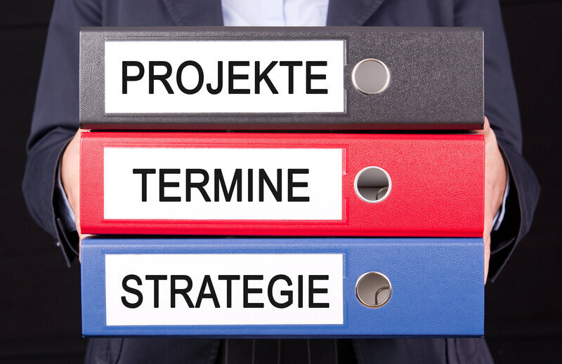 Drei Aktenordner: "PROJEKTE", "TERMINE", "STRATEGIE", von Person gehalten.