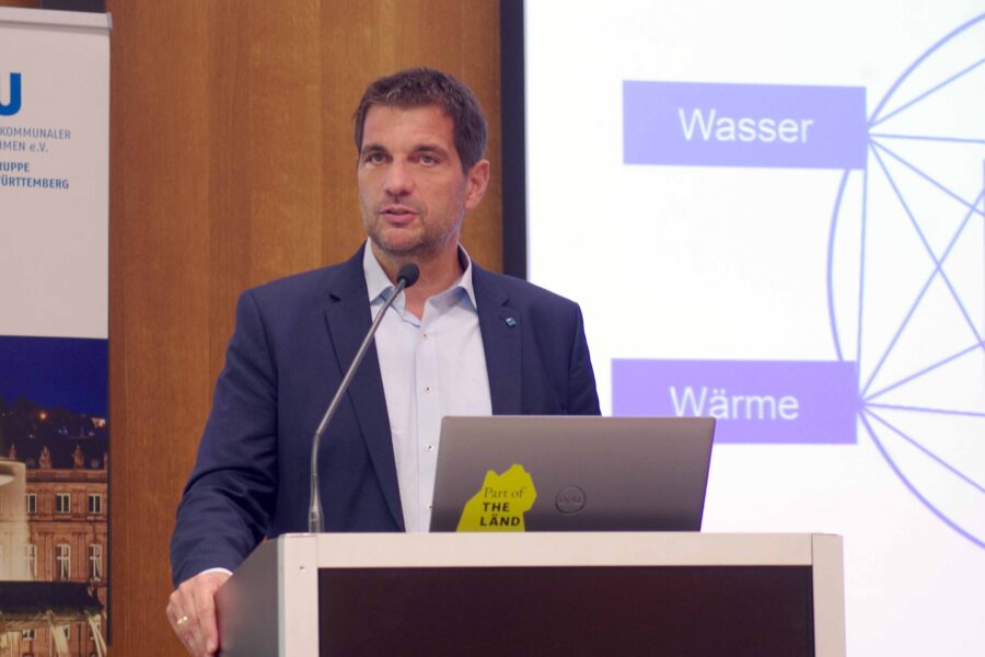 Ein Mann spricht an einem Pult mit Laptop. Text: „Wasser“, „Wärme“.