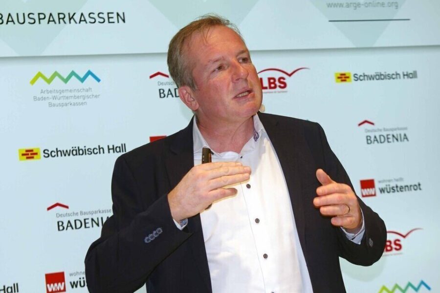 Ein Mann gestikuliert vor einer Wand mit Logos von Bausparkassen.