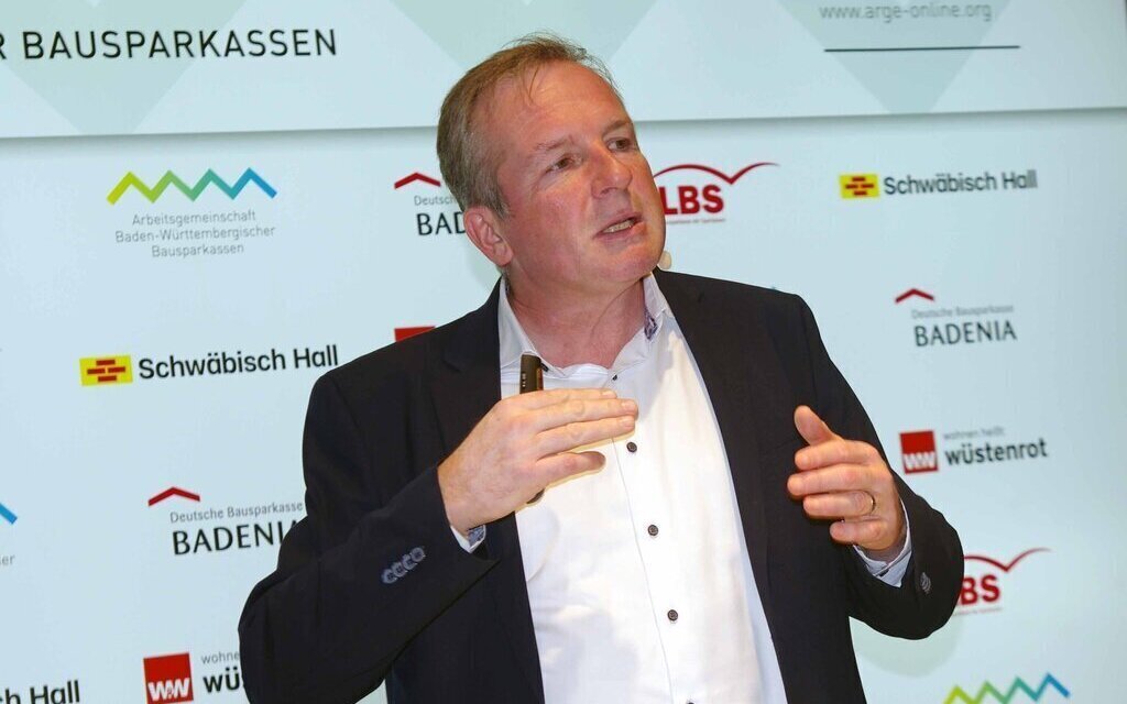 Ein Mann gestikuliert vor einer Wand mit Logos von Bausparkassen.