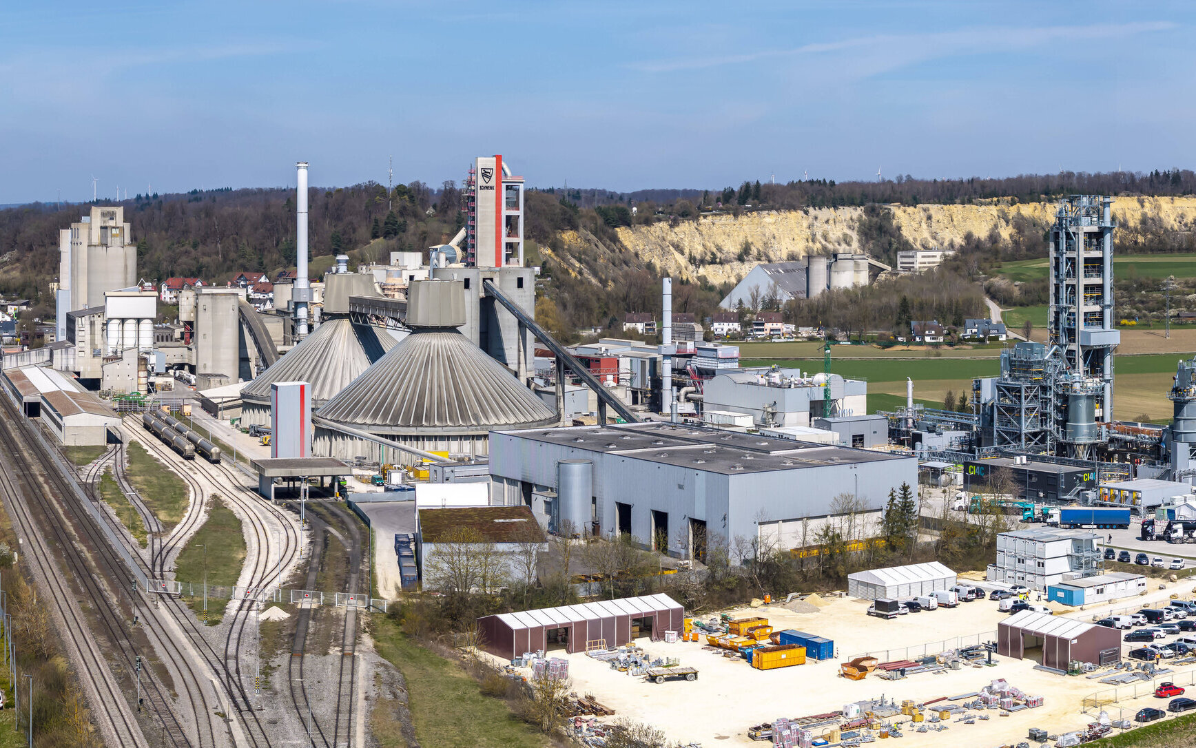 Industrieanlage mit Fabrikgebäuden, Gleisen und Lagerbereich im Vordergrund.