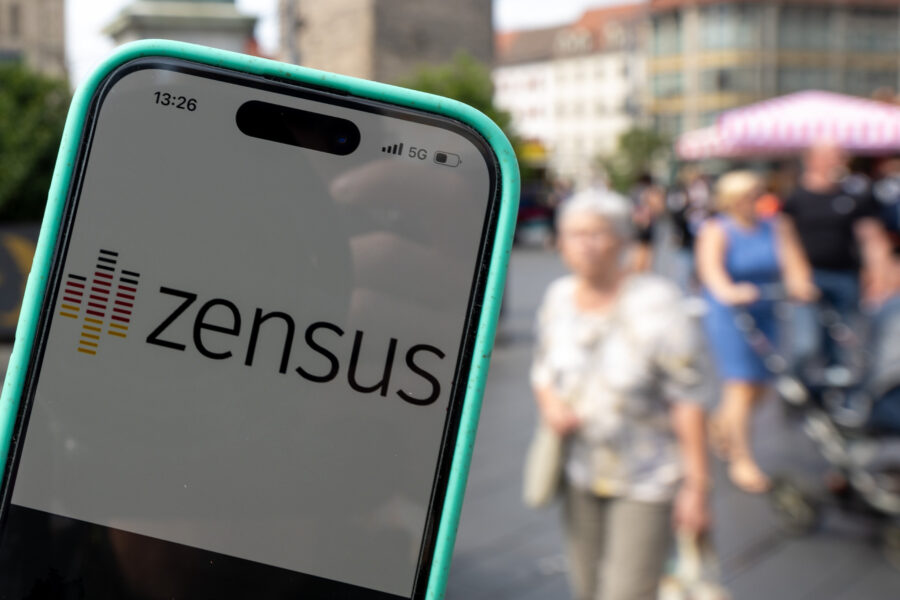Smartphone zeigt "Zensus"-App, unscharfe Passanten im Hintergrund auf Stadtstraße.